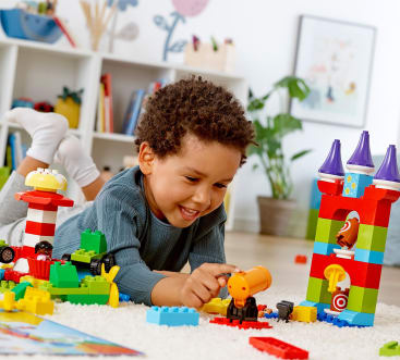 Ein kleines Kind liegt auf einem weißen Teppich und lächelt, während es mit bunten Plastikbausteinen und Spielzeugtieren spielt. Im hellen Hintergrund sind Bücherregale, Pflanzen und Wandkunst zu sehen.
