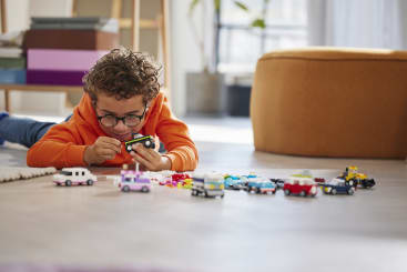 Ein Kind in einem orangefarbenen Kapuzenpulli liegt auf dem Boden und spielt mit Spielzeugautos. Mehrere bunte Spielzeugautos sind vor ihm auf einem hellen Boden in einem hellen Raum aufgereiht.