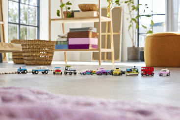 In einem hellen Wohnzimmer mit Pflanzen, Regalen und großen Fenstern im Hintergrund ist eine Reihe kleiner Spielzeugfahrzeuge, darunter Autos, ein Bus und ein Lastwagen, auf einem hellen Boden aufgereiht.