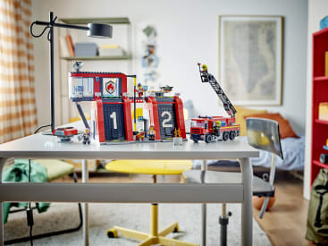 Ein LEGO Feuerwehrset mit Fahrzeugen und Figuren ist auf einem weißen Schreibtisch in einem gut beleuchteten Raum mit einem Bett, einem gelben Stuhl, einer Lampe und Regalen im Hintergrund aufgebaut.