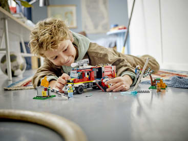 Ein kleines Kind mit blondem Haar liegt auf dem Boden und lächelt, während es mit einem LEGO-Feuerwehrauto-Set und Minifiguren spielt. Die Szene enthält zusammengesetzte LEGO Teile und eine Spielzeug-Feuerszene auf einem grauen Boden in einem Innenraum.