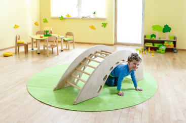 Ein kleines Kind krabbelt unter einem hölzernen Kletterbogen auf einem runden grünen Teppich in einem hellen, aufgeräumten Spielzimmer mit Holzmöbeln, Spielzeug und Dekorationen an den Wänden und in den Regalen.