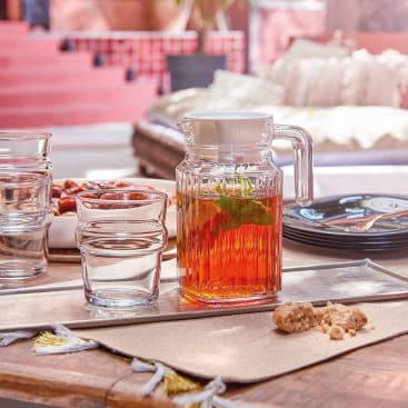 Ein mit Eistee und Minze gefüllter Glaskrug steht auf einem Tisch neben einem Glas, Tellern, Besteck, einer Schale mit Datteln und zwei Stücken braunem Zucker. Im Hintergrund sind ein Kissen und rote Stufen zu sehen.