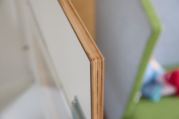 Nahaufnahme der Kante einer Sperrholzplatte, die mehrere Schichten dünner, miteinander verleimter Holzfurniere zeigt. Im verschwommenen Hintergrund sind ein grünes Objekt und einige unscharfe bunte Gegenstände zu sehen.