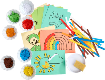 Buntes Bastelpapier mit skizzierten Mustern, Schalen mit farbigen Papierstücken, gewellte Papierstreifen, ein kleiner Schwamm und eine weiße Schale werden auf einer weißen Fläche für eine Bastelaktivität für Kinder angeordnet.