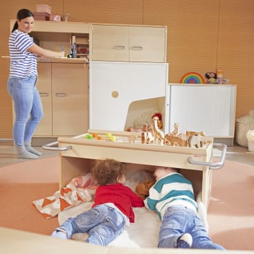 Zwei kleine Kinder liegen unter einem hölzernen Spieltisch, auf dem Tierfiguren stehen, während ein Erwachsener in einem hellen, aufgeräumten Spielzimmer neben einem Schrank steht.
