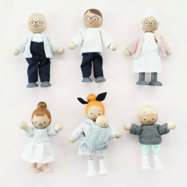 Sechs Holzpuppen sind in zwei Reihen vor einem hellen Hintergrund angeordnet. Die obere Reihe zeigt drei Erwachsene in verschiedenen Kostümen, während die untere Reihe drei Kinderpuppen zeigt, eine davon mit einem Baby im Tragetuch. Alle haben aufgemalte Gesichtszüge.