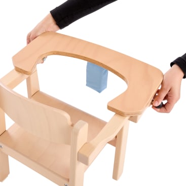 Eine Person befestigt ein Holztablett mit einem U-förmigen Ausschnitt an einem hellen Holzstuhl. Der Stuhl hat einen blauen Stoffgurt und ist für ein Kind gedacht.