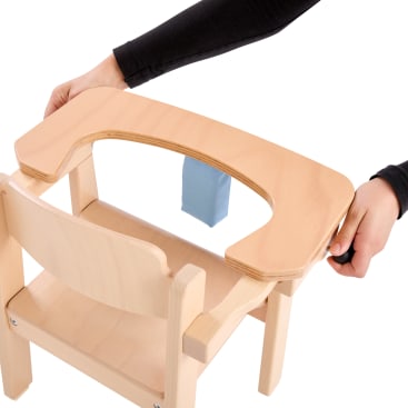Eine Person befestigt ein Holztablett mit einem blauen Gurt an einem kleinen Holzstuhl und baut wahrscheinlich einen Hochstuhl oder eine Sitzerhöhung für Kinder auf weißem Hintergrund zusammen.