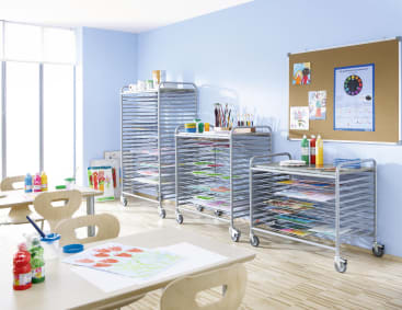 Ein Klassenzimmer mit drei Metallständern zum Trocknen von Kunstwerken, darauf Kunstzubehör, Holztische und -stühle, Farbflaschen und eine Korkwand mit Postern und Zeichnungen an einer hellblauen Wand.