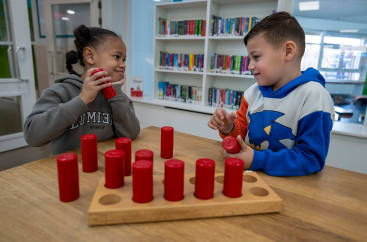 Zwei Kinder sitzen an einem Tisch und spielen mit roten zylindrischen Gegenständen, die sie in ein Holzbrett legen. Im Hintergrund sind Bücherregale zu sehen, und beide Kinder scheinen sich auf ihre Tätigkeit zu konzentrieren.