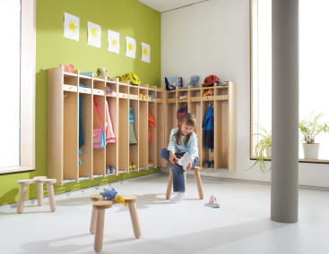 Ein junges Mädchen sitzt auf einem Holzschemel und zieht sich in einer hellen, gut organisierten Garderobe mit Fächern für Mäntel, Taschen und Helme die Schuhe an. Die Wände sind grün und mit Kinderzeichnungen von gelben Sonnen verziert.