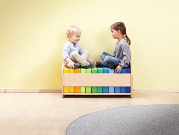 Zwei Kinder sitzen auf einer Holzbank mit bunten Blöcken darunter, schauen sich an und scheinen sich zu unterhalten. Der Hintergrund besteht aus einer gelben Wand und einem hellen Holzboden mit einem Teil eines grauen Teppichs.