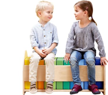 Ein Junge und ein Mädchen sitzen sich auf einer Holzbank gegenüber, unter der bunte Blöcke liegen. Der Junge trägt ein hellblaues Hemd und eine beigefarbene Hose, während das Mädchen ein graues Oberteil und eine Jeans mit Herzaufnähern an den Knien trägt.