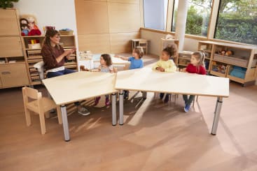 Eine Lehrerin sitzt mit vier kleinen Kindern an einem Tisch in einem Klassenzimmer mit Holzmöbeln und Regalen mit Spielzeug und Büchern. Die Lehrerin hält ein Tablet in der Hand, und die Kinder sitzen und schauen in ihre Richtung.