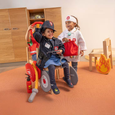 Zwei kleine Kinder spielen Verkleiden; eines trägt ein Feuerwehrkostüm und sitzt auf einem Spielzeug-Feuerwehrauto, während das andere ein Arztkostüm trägt und daneben steht. Sie befinden sich in einem Spielzimmer mit Holzmöbeln und einer Spielzeug-Feuerwehrrequisite.