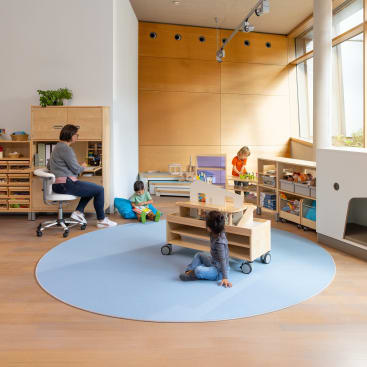 Eine Lehrerin sitzt an einem Schreibtisch, während drei kleine Kinder in einem hellen Klassenzimmer mit Holzmöbeln und großen Fenstern auf dem Boden und in Regalen spielen und lesen.