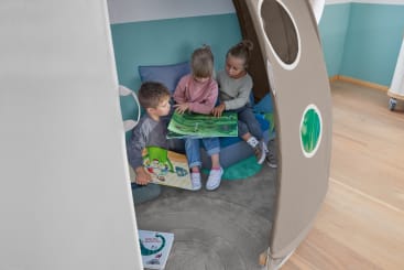 Drei kleine Kinder sitzen zusammen in einem braunen Spielzelt auf einem grauen, runden Teppich und betrachten Bilderbücher. Das Zelt hat runde Fenster und Kissen, und der Raum hat einen hellen Holzboden und grün-blaue Wände.