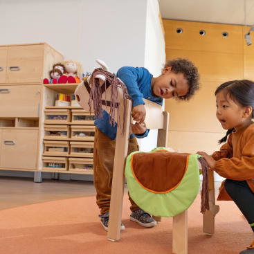 Zwei kleine Kinder spielen mit hölzernen Steckenpferden in einem Haus. Sie scheinen beschäftigt zu sein, wobei eines steht und das Pferd einstellt und das andere in der Nähe hockt. Im Hintergrund befinden sich Regale mit Spielzeug und Puppen.