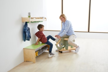 Ein Erwachsener hilft einem Kind in einem hellen Raum beim Anziehen der Schuhe. Das Kind sitzt auf einer Bank mit einem grünen Kissen, und auf einem Regal darüber hängen ein Mantel und ein Rucksack. Der Erwachsene kniet auf einem Hocker in Form eines Elefanten.