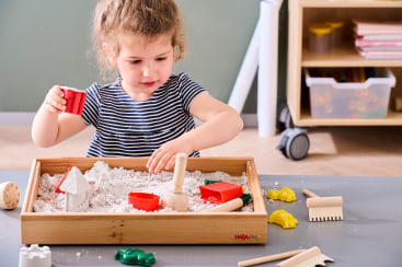Ein kleines Kind mit lockigem Haar spielt mit Sand in einer hölzernen Sinnesbox und verwendet rote Formen und Holzwerkzeuge. Auf dem Tisch daneben liegen verschiedene Spielzeugformen und Werkzeuge. Im Hintergrund befindet sich ein Regal mit Aufbewahrungsbehältern.