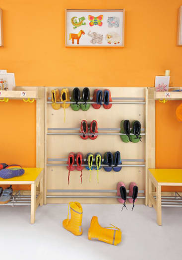 In einem hölzernen Schuhregal stehen sieben Paar Kinderschuhe in verschiedenen Farben. Zwei gelbe Regenstiefel stehen auf dem Boden vor dem Regal. Die Wand hinter dem Regal ist orangefarben, und über dem Regal hängen Kunstwerke von Kindern.