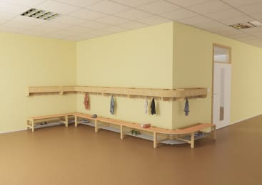 Ein sauberer, leerer Umkleideraum mit hellgelben Wänden, Holzbänken, Haken mit ein paar hängenden Kleidungsstücken und Regalen für Schuhe. Der Raum hat einen braunen Boden, eine Decke mit Leuchtstoffröhren und eine Tür mit Glasscheiben.