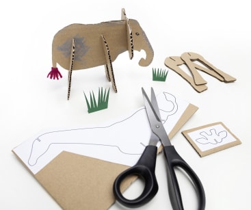 Ein Elefant aus Pappe wird mit zusätzlichen Pappteilen, Papierschablonen, grünem Papiergras und einer Schere auf einer weißen Fläche zusammengebaut.
