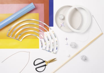 Eine Reihe von Bastelmaterialien, die auf einer weißen Fläche ausgelegt sind, darunter farbige Papierbögen, Scheren, Klebeband, ein Metalldraht, ein Holzdübel, ein holografischer regenbogenförmiger Ausschnitt, Pompons, Faden und sternförmige Verzierungen.