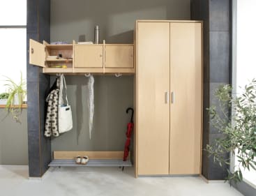 Eine hölzerne Garderobe und ein Schrank an einer grauen Wand, mit offenen und geschlossenen Fächern, hängenden Schals, Taschen, einem roten Regenschirm, weißen Schuhen und einem Gegenstand aus rostfreiem Stahl obenauf. Auf beiden Seiten sind Pflanzen aufgestellt.