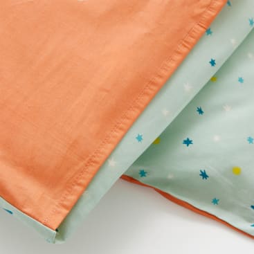 Zwei Stoffstücke: eines ist einfarbig orange mit sichtbaren Nähten, das andere ist hellblau-grün mit einem Muster aus kleinen, bunten Sternen. Die Stoffe überlappen sich auf einer ebenen Fläche.