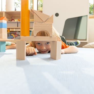 Ein kleines Kind liegt auf dem Boden und schaut durch eine Struktur aus Holzbausteinen. Der Hintergrund zeigt ein Spielzimmer mit weiteren Bauklötzen und natürlichem Licht, das durch die Fenster fällt.