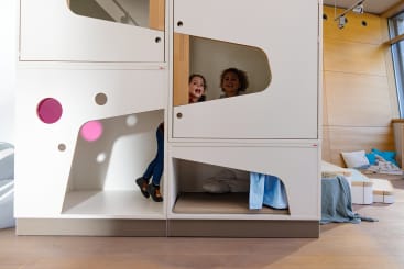 Zwei Kinder spielen in einem modernen, weißen Indoor-Spielgerät mit geometrischen Aussparungen. Ein Kind schaut heraus und lächelt, während das andere neben ihm steht. Der Raum hat Holzfußböden und Licht, das aus den Fenstern einfällt.