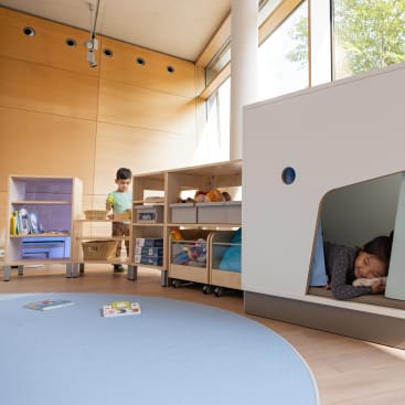Ein kleines Kind steht in einem hellen Raum mit Holzwänden neben Regalen mit Spielzeug und Büchern, während ein anderes Kind in einer kleinen, eingebauten Ecke schläft. Auf dem Boden liegt ein runder blauer Teppich, auf dem zwei Bücher liegen.