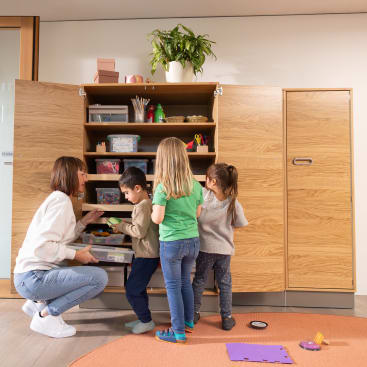 Ein Erwachsener und drei kleine Kinder stehen vor einem offenen Holzschrank mit Regalen mit Kunstzubehör. Der Erwachsene kniet nieder und hilft den Kindern bei der Auswahl der Gegenstände. Die Szene spielt sich in einem hellen, aufgeräumten Raum ab.