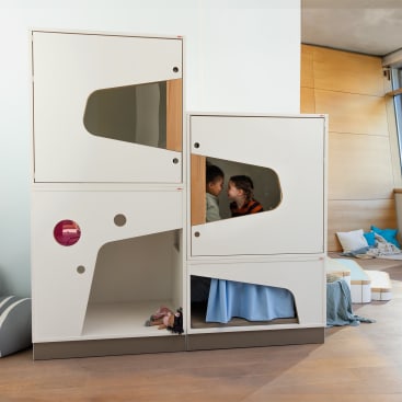 Zwei Kinder sitzen in einer modernen modularen Spielstruktur mit geometrischen Ausschnitten in einem hellen Raum mit Holzboden und hellen Wänden; im Hintergrund befinden sich weiche Sitzmöbel und Kissen.