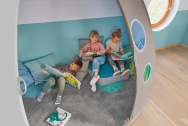 Drei kleine Kinder sitzen und liegen auf Kissen und lesen Bücher in einem gemütlichen, modernen Spielzimmer mit tealfarbenem und grauem Dekor, runden Fenstern und einem hellen Holzboden.