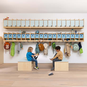 Zwei kleine Kinder sitzen auf Holzbänken in einem Klassenzimmer oder einer Kindertagesstätte. Hinter ihnen stehen Regale mit blauen Aufbewahrungsboxen, Fächern und Haken, an denen verschiedene Rucksäcke und Jacken hängen. Der Boden ist aus hellem Holz.