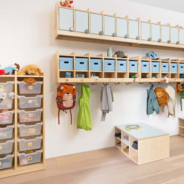 Ein ordentlicher Aufbewahrungsbereich im Klassenzimmer mit Fächern, Behältern und Haken. Rucksäcke, Jacken und Stofftiere sind entlang der Wand aufgereiht. Auf der linken Seite sind durchsichtige Plastikschubladen gestapelt, und unter den Fächern befindet sich eine kleine Bank mit Regalen.