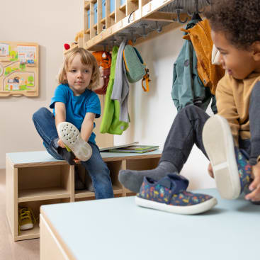 Zwei Kinder sitzen auf Bänken in einer Umkleidekabine und ziehen sich die Schuhe an. Hinter ihnen hängen Mäntel und Rucksäcke an Haken, und im Hintergrund ist ein an der Wand befestigtes Spielbrett zu sehen.