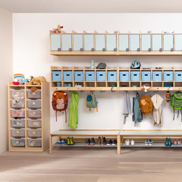 Eine aufgeräumte Garderobe mit Fächern, Behältern und Haken für die Aufbewahrung. Rucksäcke, Jacken, Schuhe und Hüte werden in Regalen aufbewahrt, Kuscheltiere und Spielzeug in übersichtlichen Schubladen auf der linken Seite. Unter den Ablageflächen befinden sich Bänke.