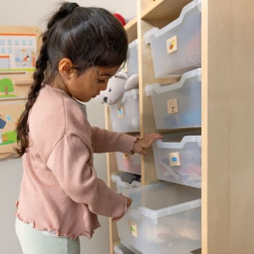 Ein junges Mädchen mit langen dunklen Haaren und einem rosa Pullover öffnet eine Plastikschublade in einem Holzregal. Das Regal enthält mehrere Schubladen, die jeweils mit einem Aufkleber versehen sind. Die Szene scheint in einem Kinderzimmer zu spielen.
