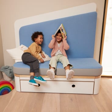 Zwei Kinder sitzen auf einer blau-weißen Bank in einem Haus. Ein Kind hält sich ein Buch über den Kopf und lächelt, während das andere lacht. Eine Decke und ein buntes Regenbogenspielzeug aus Holz liegen in der Nähe auf dem Boden.