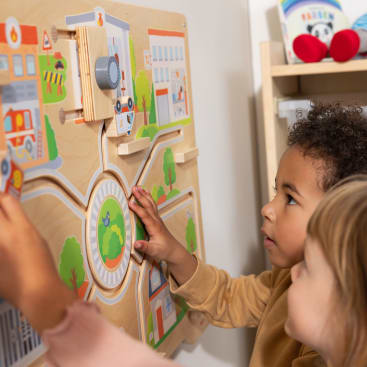 Zwei kleine Kinder stehen nebeneinander und interagieren mit einer hölzernen Wandtafel, die bunte Illustrationen von Gebäuden, Straßen und Fahrzeugen zeigt.