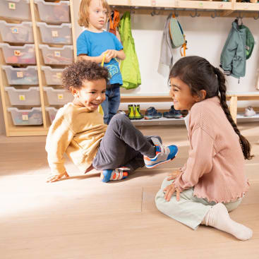 Zwei Kinder sitzen in einem Klassenzimmer auf dem Boden und lächeln sich gegenseitig an, während ein drittes Kind im Hintergrund neben Regalen mit Behältern und Hängetaschen steht. Der Raum wirkt hell und aufgeräumt.