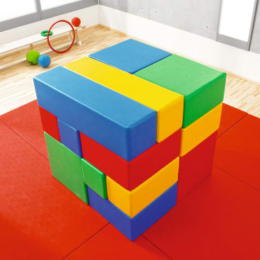 Schaumstoffblöcke in den Farben Rot, Blau, Gelb und Grün werden auf einer roten Matte in einem Spielzimmer zu einem Würfel gestapelt. Im Hintergrund liegen einige Ringe und Bälle auf einem Holzboden.