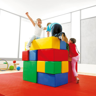 Drei Kinder klettern und spielen auf großen, bunten Schaumstoffblöcken, die in einem hellen Spielzimmer mit roten Matten und Turngeräten im Hintergrund gestapelt sind. Ein Kind sitzt mit erhobenem Arm obenauf.