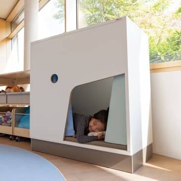 Ein kleines Kind liegt in einer kleinen, modernen, weißen Schlafkabine mit offener Seite in einem hellen Raum mit großen Fenstern und einfallendem Sonnenlicht. Das Kind scheint zu ruhen oder zu schlafen.
