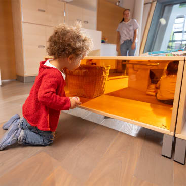 Ein kleines Kind in einem roten Pullover kniet auf einem Holzboden und blickt in einen niedrigen, gelb getönten Spiegelschrank. Ein Erwachsener steht im Hintergrund neben Regalen und einem Fenster.