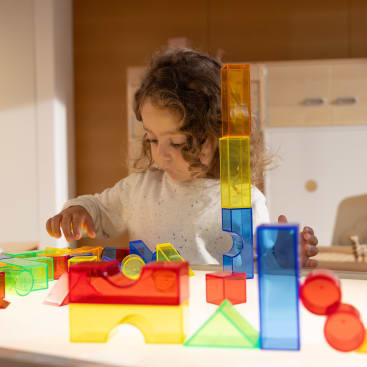 Ein kleines Kind mit lockigem Haar spielt mit bunten, durchsichtigen Plastikbausteinen auf einem Tisch in einem gut beleuchteten Raum. Das Kind konzentriert sich auf das Stapeln und Anordnen der Blöcke.